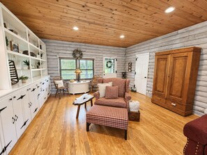 A cozy cabin interior with a wooden ceiling, a plaid sofa, a white cabinet with drawers, and a large wooden wardrobe.
