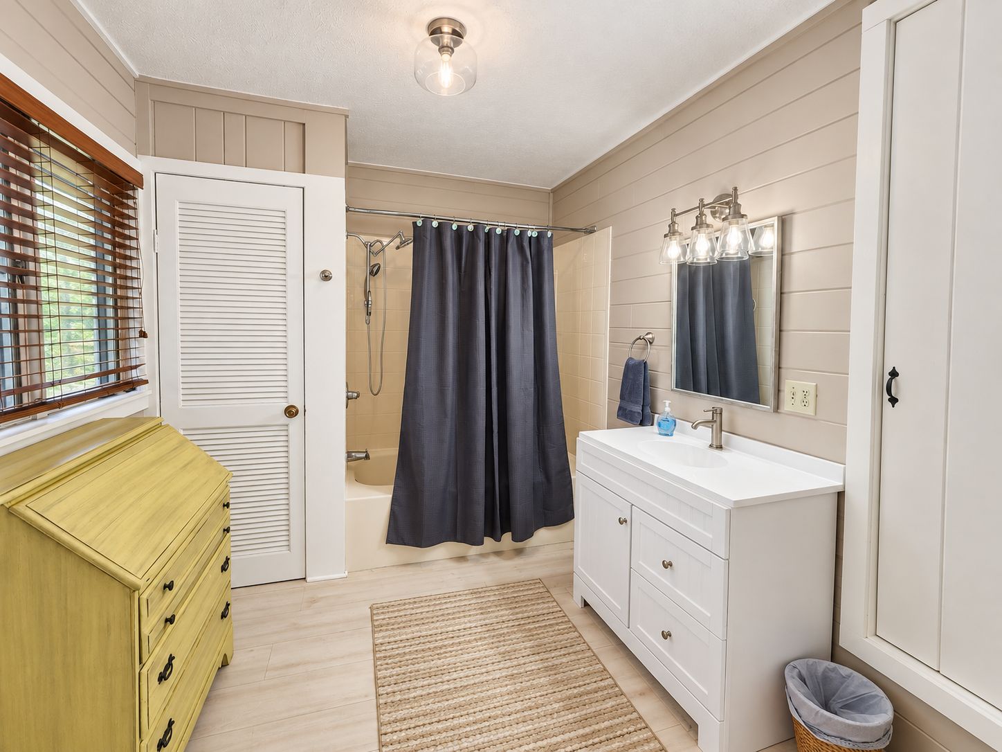 Een badkamer met een douche, een wastafel met spiegel, een opbergmeubel en een raam met zonwering.