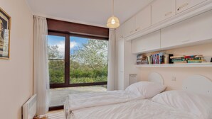 A bedroom with a large window, a bed, a bedside table, and a shelf with books. - ZeeZonZand A-GV01 + P* (Koksijde)