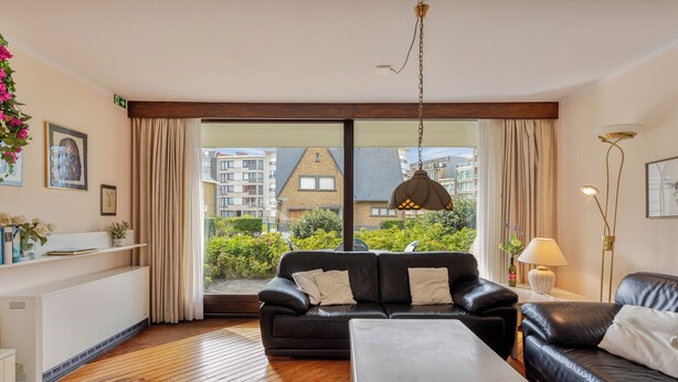 A modern living room with a black leather sofa, a white coffee table, and a view of residential buildings through large windows. - ZeeZonZand A-GV01 + P* (Koksijde)