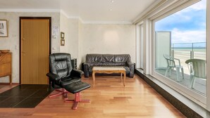 Un salon moderne avec un plancher en bois, un fauteuil en cuir noir, une table basse en bois, un canapé gris et une grande fenêtre donnant sur la plage.