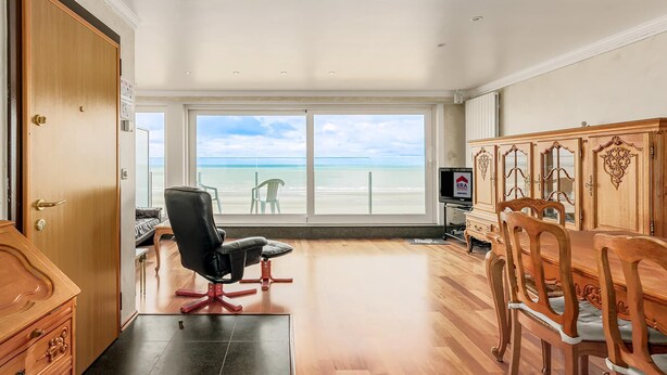 Une pièce spacieuse comprenant un ensemble de salle à manger en bois, un fauteuil de bureau noir et un téléviseur. Une grande fenêtre offre une vue sur la plage.