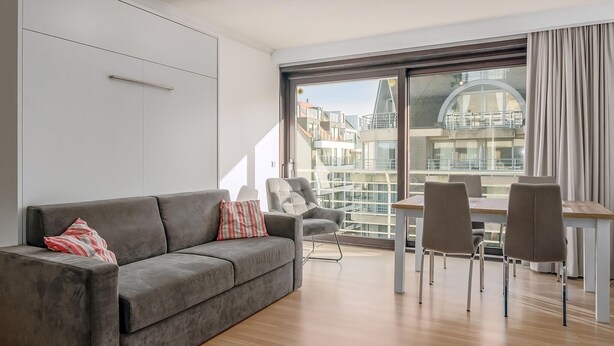 Una sala de estar moderna con un sofá gris, una mesita con sillas y un ventanal con vistas a edificios.