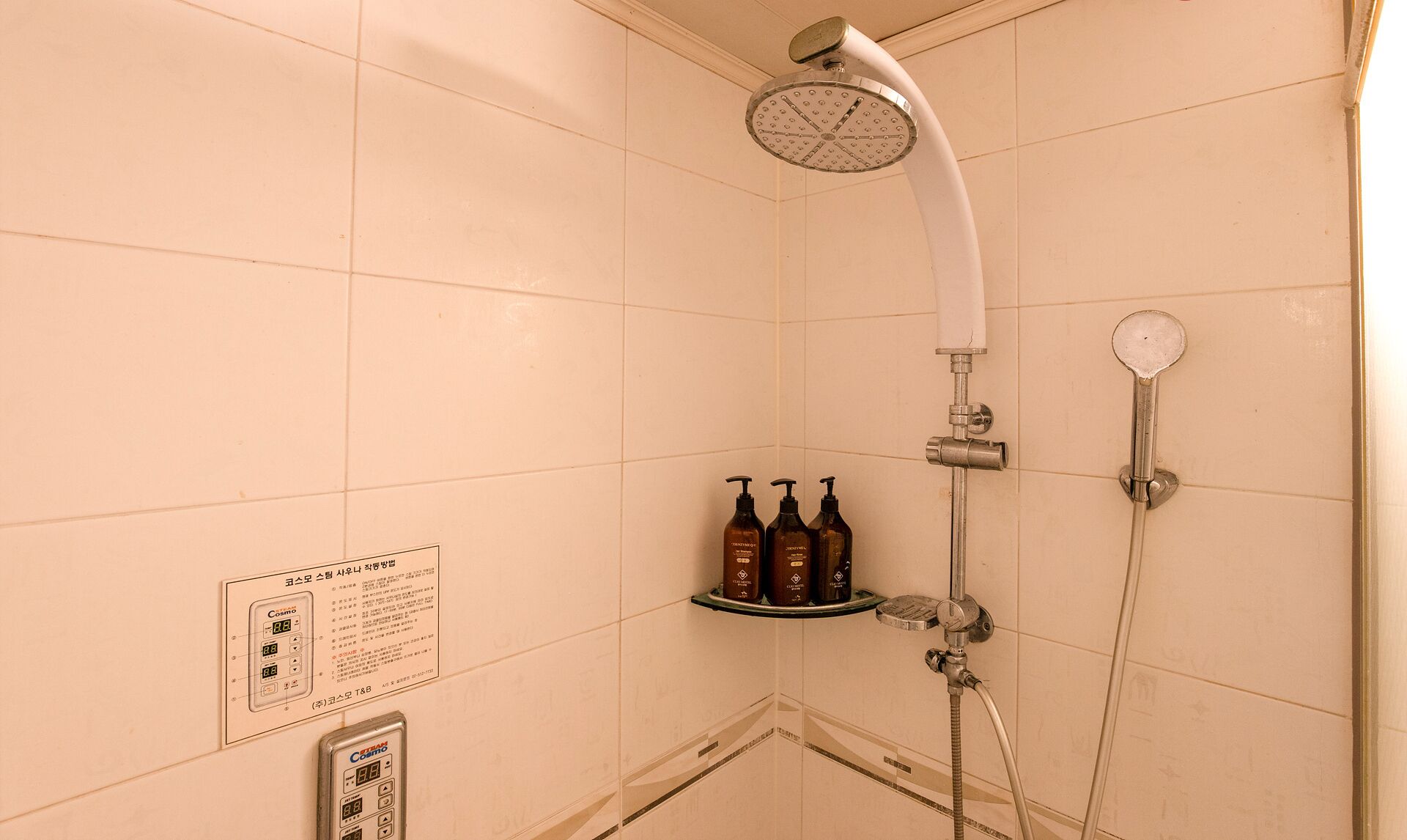 A shower with a handheld showerhead, a wall-mounted faucet, and a handheld shower attachment. There are three bottles of shampoo on a shelf.