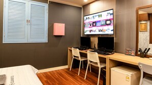A hotel room with a wooden desk, two white chairs, a television mounted on the wall displaying various channels, a small pink box on the wall, and a bed with a white cover. - CLIO MOTEL (Incheon)