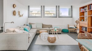 A modern living room with a beige sofa, white and wooden side tables, a bookshelf, and a view through large windows.