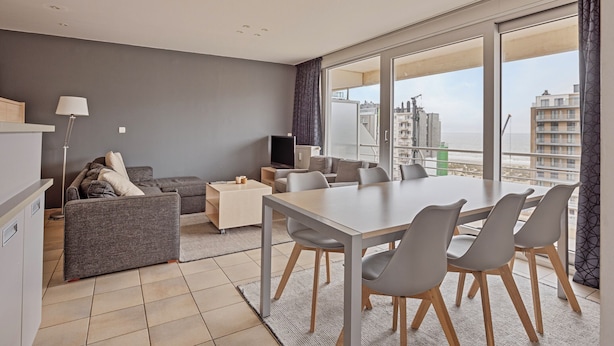 A modern living room with a grey sofa, a dining table with chairs, a TV, and a large window with a view of buildings.