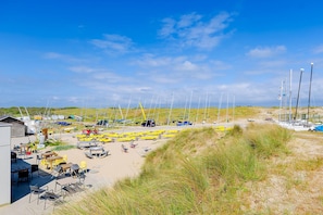 A beachside area with multiple sailboats, a sandy beach, and a small building with outdoor seating.