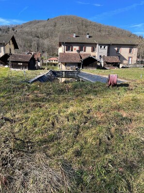 Ländliche Gegend mit traditionellen Häusern, einer grünen Wiese und einer Hügelkulisse im Hintergrund.