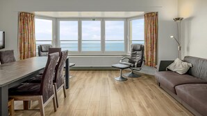 A modern living room with a large bay window, a dining table with chairs, a sofa, and a floor lamp. - 2 bedroom accommodation in De Panne (De Panne)