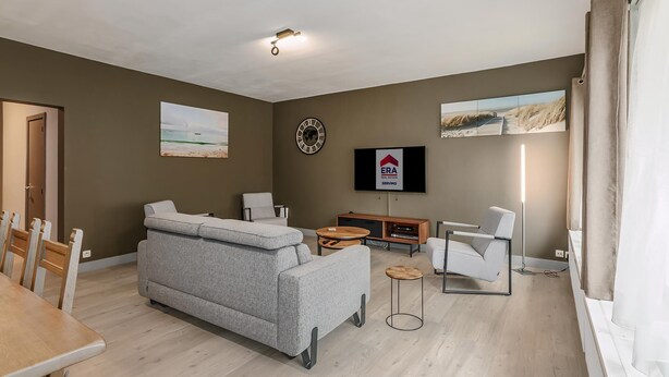 Un salon moderne avec un canapé gris, des fauteuils blancs, une table à manger et un téléviseur à écran plat fixé au mur.
