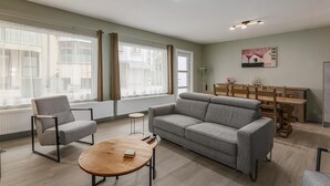 Un salon moderne avec un canapé gris, une table basse ronde en bois et un fauteuil gris. On y trouve également un coin repas avec une table et des chaises en bois.