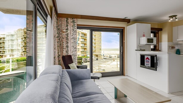 A modern apartment with a grey sofa, a dining table, a kitchen with a microwave, and a large sliding glass door to a balcony with a view of high-rise buildings and a beach. - 2 bedroom accommodation in Sint Idesbald (Sint Idesbald)