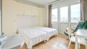 A bedroom with two single beds, a white sink, a green chair, and a view of the city through large windows.