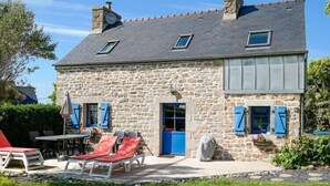 A stone house with a sloped roof, a small porch with red lounge chairs, and a blue door.