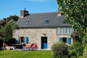 Een stenen huis met schuin dak, twee ramen met luiken, een blauwe deur en een klein terras met een tafel en stoelen.