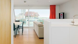 A modern kitchen with a large island, a dining area with a table and chairs, and a view of buildings through large windows. - 2 bedroom accommodation in Oostduinkerke (Oostduinkerke)