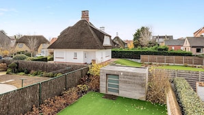 Une maison avec un toit de chaume, un petit garage attenant et un jardin bien entretenu avec une pelouse et des haies.