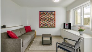 Un salon moderne avec un canapé gris, des coussins rouges, une petite table, un fauteuil, une courtepointe colorée accrochée au mur et une fenêtre donnant sur un paysage agréable.