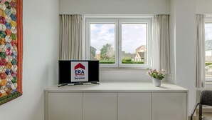 Une chambre moderne avec une grande fenêtre, un téléviseur à écran plat posé sur un meuble blanc, une couverture aux couleurs vives et une plante en pot.