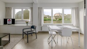 Une pièce moderne et bien éclairée, comprenant un coin repas, un téléviseur posé sur un meuble et de grandes fenêtres donnant sur le quartier.