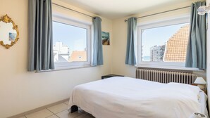 Une chambre avec deux fenêtres, un lit, un radiateur, une table de chevet et un miroir.
