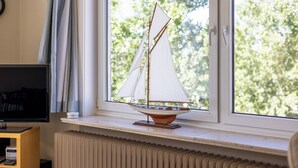 Un modèle de voilier est posé sur un rebord de fenêtre, un radiateur se trouve en dessous, et un téléviseur est installé sur un meuble à gauche.