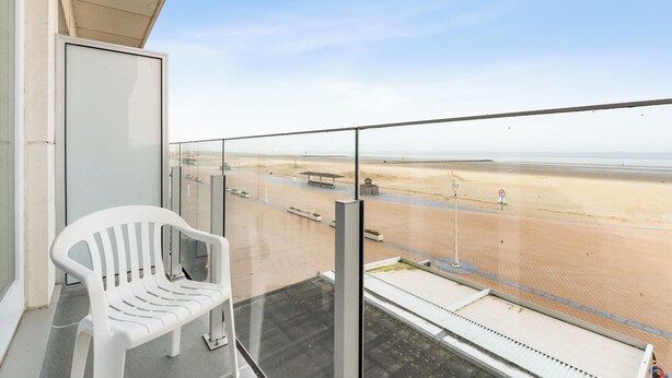 Ein Balkon mit weißem Kunststoffstuhl, einer Glastrennwand und Blick auf einen Sandstrand.