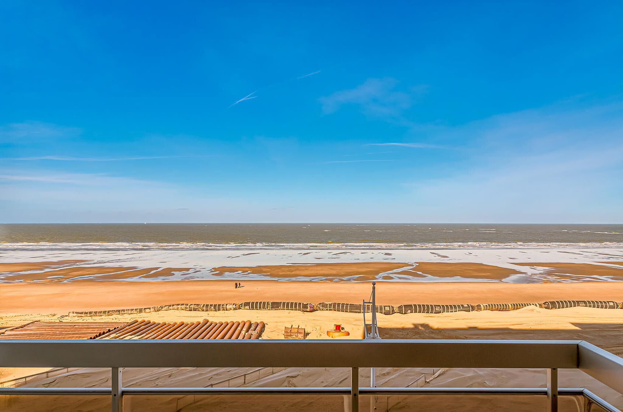 A beach view from a balcony with a clear sky, sandy beach, and a wooden deck.