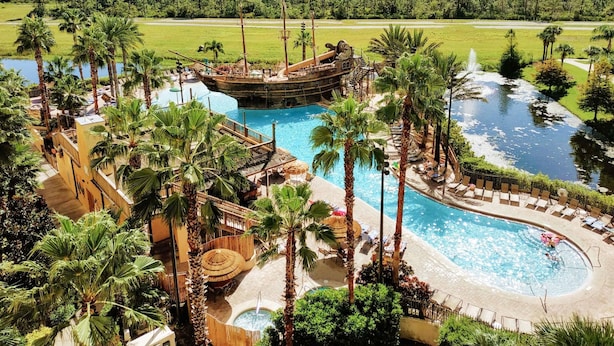 A resort with a large swimming pool, a ship-themed water slide, palm trees, and a view of the surrounding forest. - Upstay - Spacious Suite at Kid-friendly Resort (Orlando)