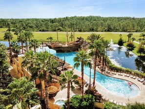 A resort with a large swimming pool, a ship-themed water slide, palm trees, and a view of the surrounding forest.