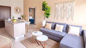 Een moderne woonkamer met een grijze bank, een klein eiland met een spoelbak en een macramé wanddecoratie.