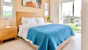 Een slaapkamer met een bed, nachtkastje, lamp en een raam met uitzicht op een stadsbeeld.