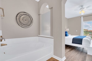 Een moderne badkamer met een ruim bad, een rond kunstwerk aan de muur en uitzicht op een slaapkamer met een bed en balkon.