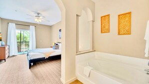 Une chambre à coucher comprenant un lit, un ventilateur de plafond, une fenêtre avec des rideaux, un balcon et une salle de bain avec une baignoire et des panneaux muraux décoratifs.
