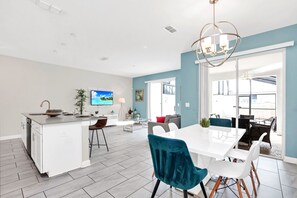 A modern kitchen with a central island, a dining area with a white table and blue chairs, and a large window providing natural light. - Upstay - Modern Home w Pool Near Disney (Kissimmee)