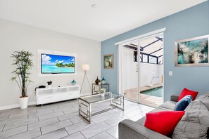 A modern living room with a flat-screen TV, a glass coffee table, a sofa with red cushions, a potted plant, and a pool area visible through glass doors. - Upstay - Modern Home w Pool Near Disney (Kissimmee)