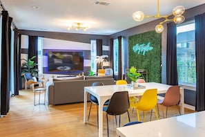 A modern living room with a sofa, dining table, and chairs, a flat-screen TV, and a green wall with a neon sign. - Hot Tub Time Machine Clubhouse DT 6 Min Drive (Austin)