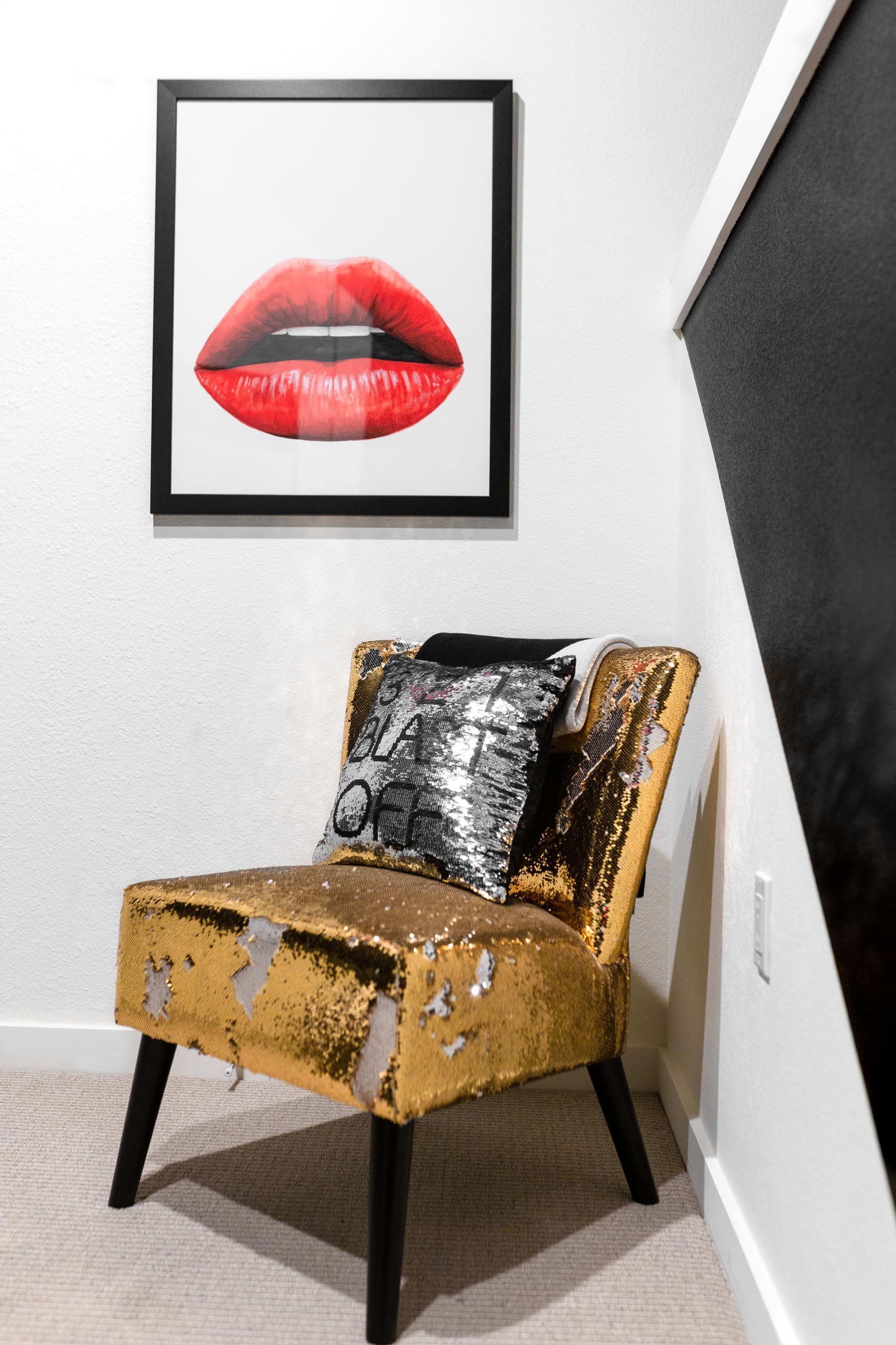 Een kamer met een lijstwerk met rode lippen, een vergweerde gouden stoel en een zwart-zilver kussen.