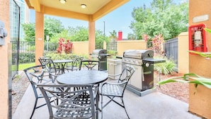 Une terrasse extérieure avec une table ronde et des chaises, un barbecue et une poubelle.