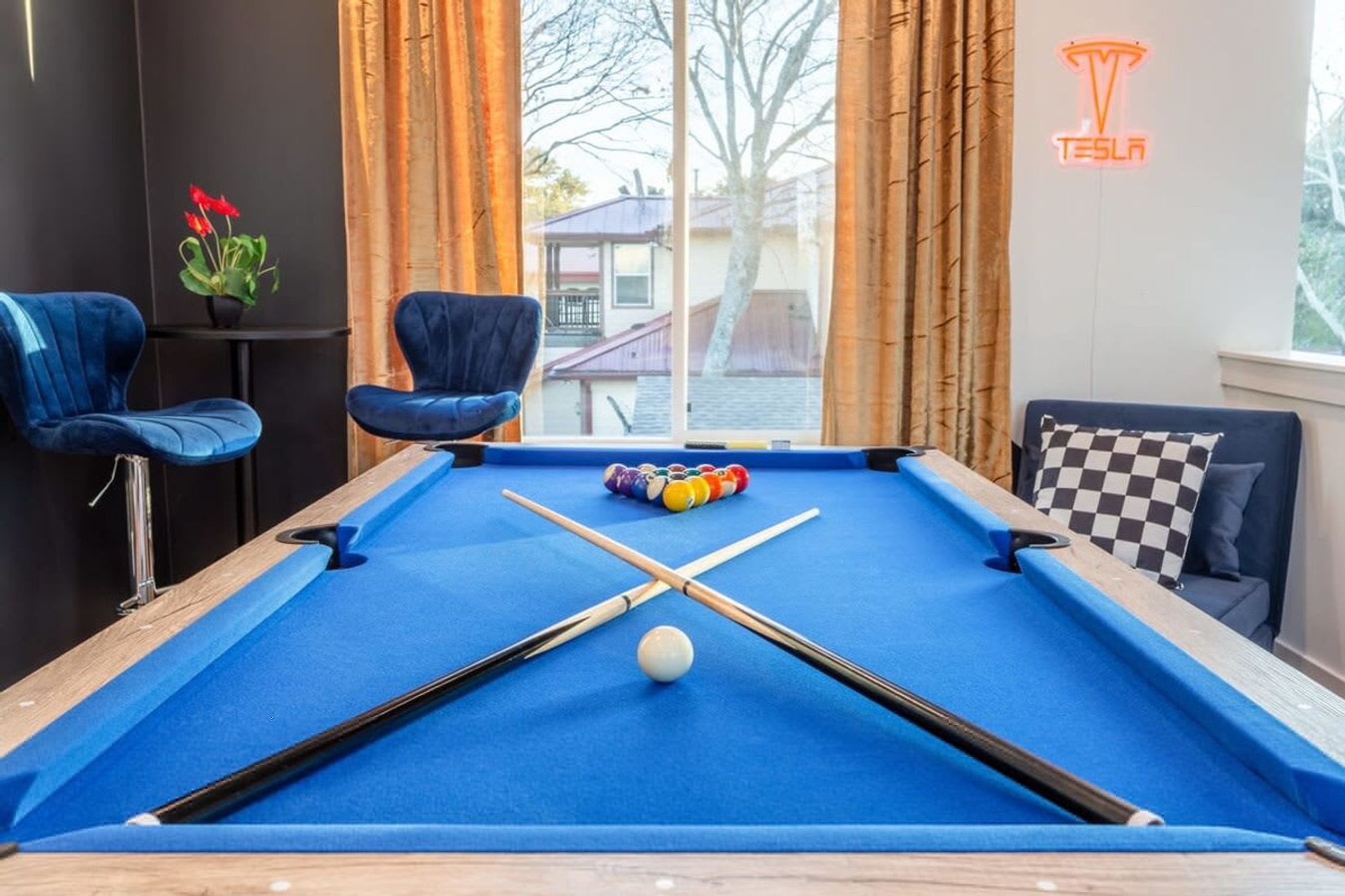 A pool table with pool cues and balls, a blue chair, a sofa with checkered cushions, and a Tesla logo on the wall.