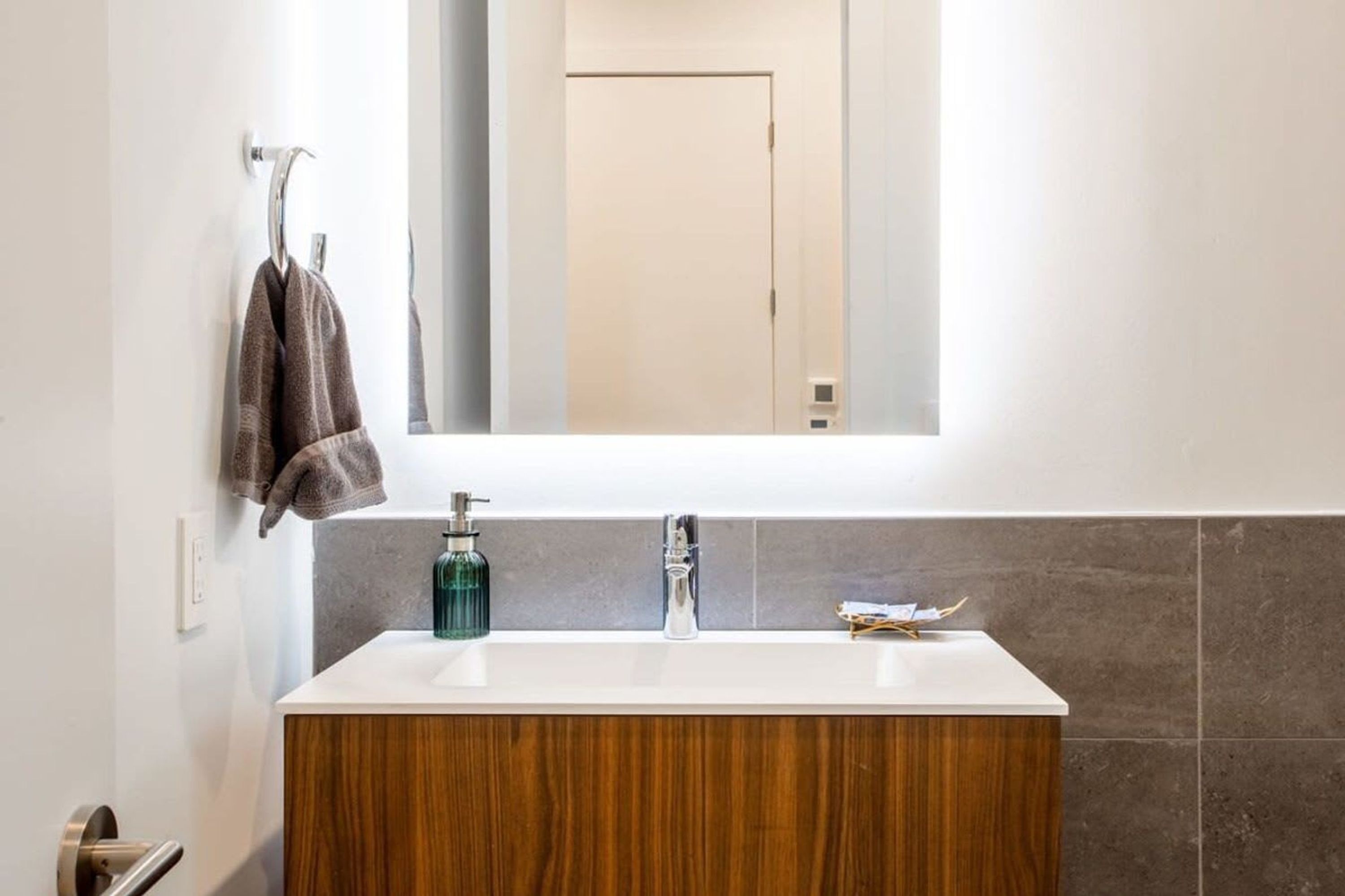 Un baño con tocador de madera, un lavabo blanco, un espejo sobre el lavabo y una toalla colgada en un gancho.