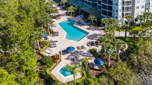 An aerial view of a resort with a large, irregularly shaped pool surrounded by lounge chairs and umbrellas, set against a backdrop of palm trees and a multi-story building.
