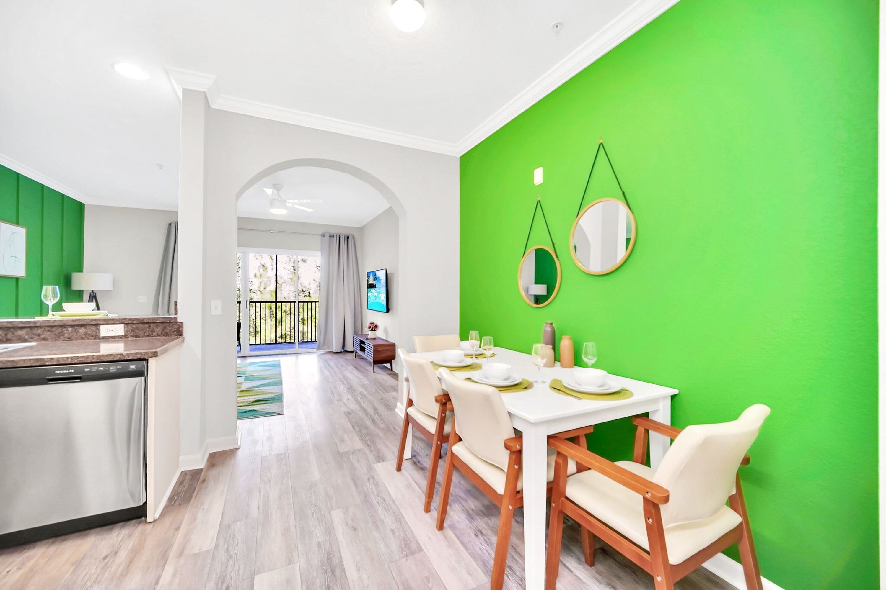 A modern kitchen with a dishwasher, a dining area with a white table and chairs, and a bright green accent wall with two round mirrors.