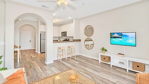 A modern living room with a TV, seating area, kitchen, and a ceiling fan.