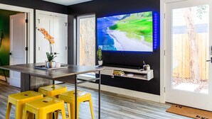 A modern kitchen with a large flat-screen TV mounted on the wall, a wooden dining table, yellow bar stools, and a view of a backyard through a glass door.