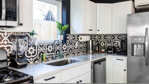 A modern kitchen with white cabinets, a stainless steel refrigerator, a built-in oven, a blender, a coffee maker, and a sink with a decorative backsplash.