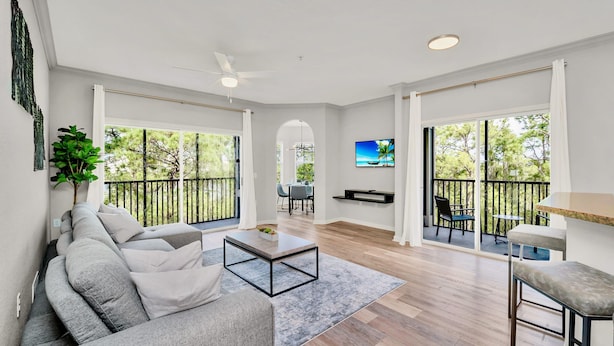 A spacious living area with a grey sofa, a coffee table, and a TV mounted on the wall. There is a balcony with a railing and a view of trees.