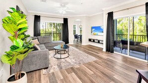 A spacious living room with a grey sofa, a coffee table, and a large plant. There is a balcony with outdoor furniture and a TV mounted on the wall. - Upstay - Modern Family Apartment w Pool Hot Tub (Winter Garden)