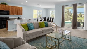 Una sala de estar moderna con un sofá color beige, una mesa de centro de vidrio y una cocina con armarios de madera y un microondas negro.